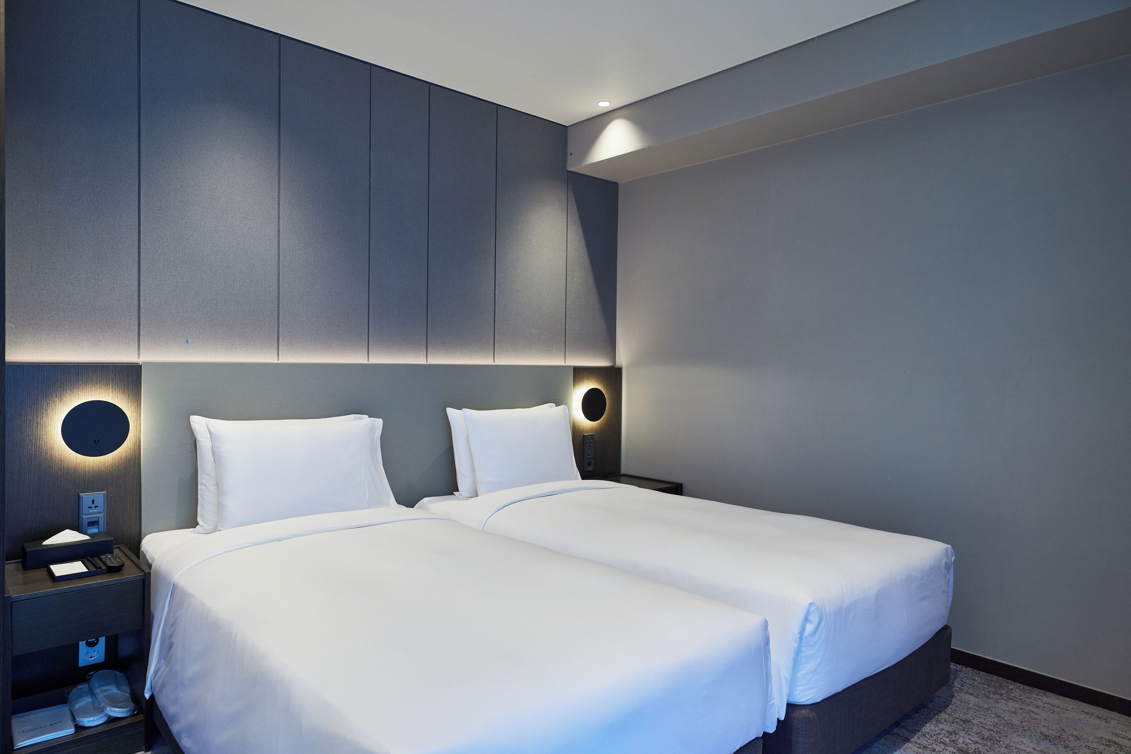 A modern hotel room with two beds, a nightstand, and a wall-mounted light fixture.