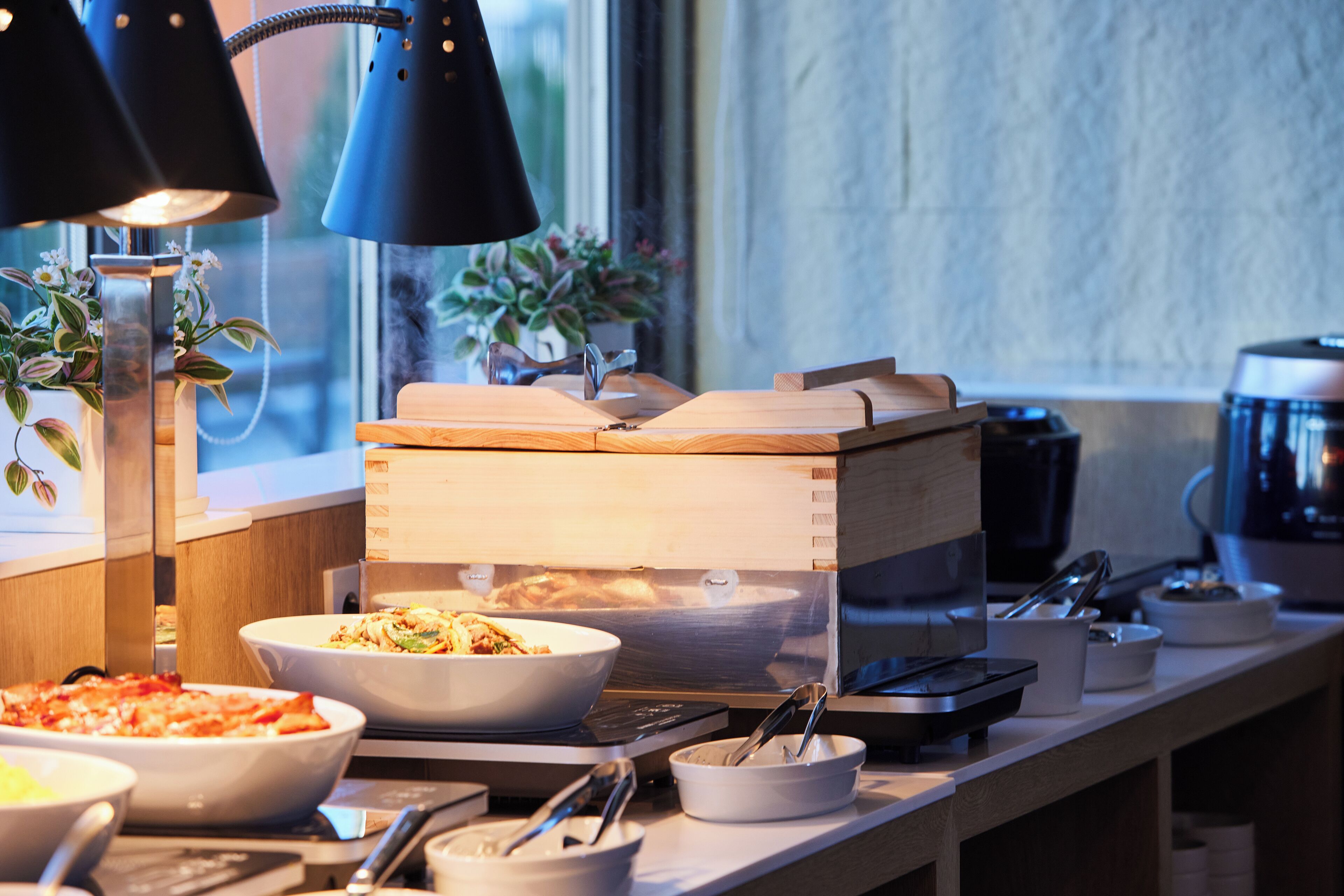 A buffet setup with various dishes, a coffee machine, and a window with plants.