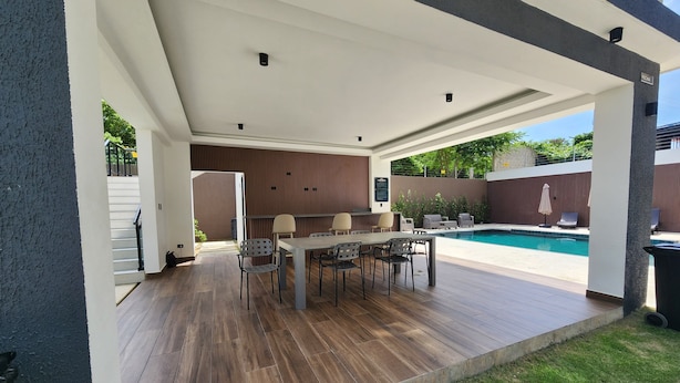 An outdoor patio area with a pool, wooden flooring, and a covered seating area with tables and chairs.