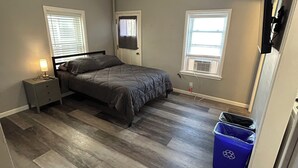 A bedroom with a bed, a ceiling fan, a nightstand, a lamp, two windows, and a trash bin. - The Loft small studio apartment on the 2nd floor.  Private, Small & Affordable (Albert Lea)