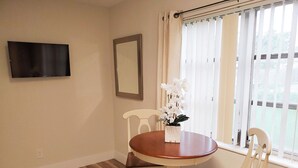 A dining area with a round table, two chairs, a vase with white flowers, a flat-screen TV on the wall, and a window with blinds. - Dania Beach 1BR Retreat - Private Entry & Self Check-In (Dania Beach)