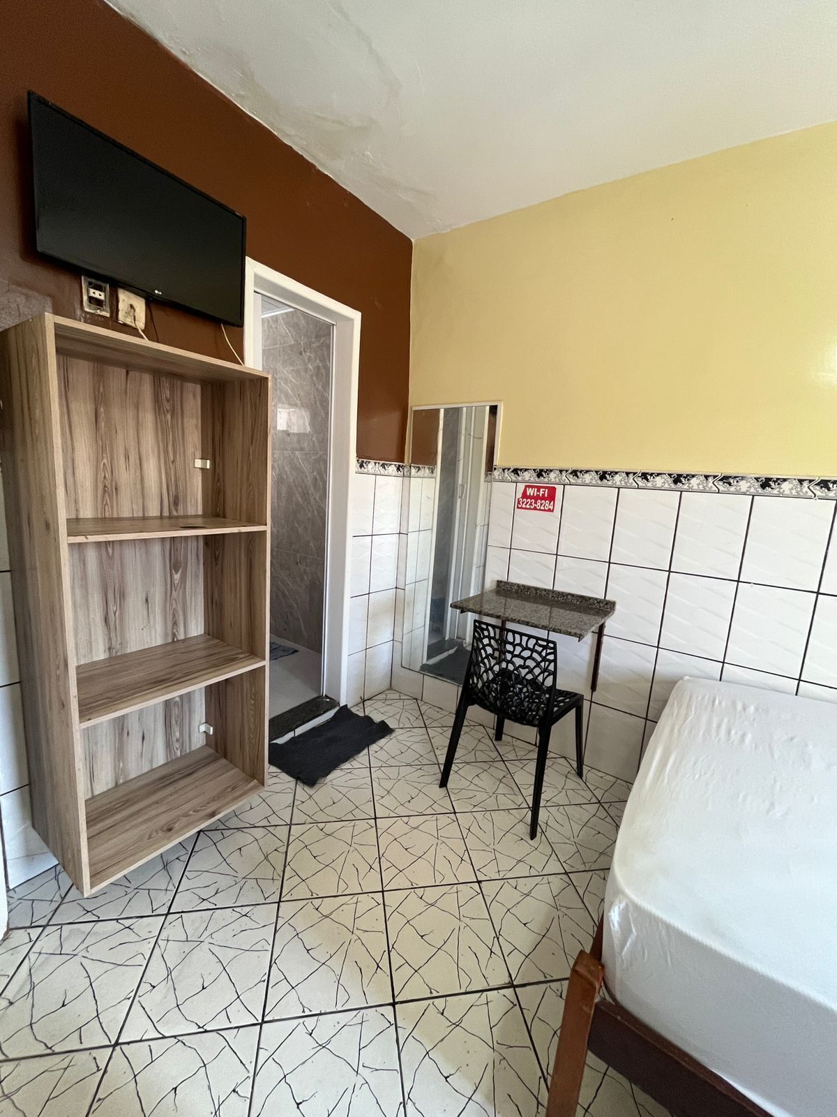 A compact room with a bed, a wooden shelf unit, a small table, and a tiled floor.