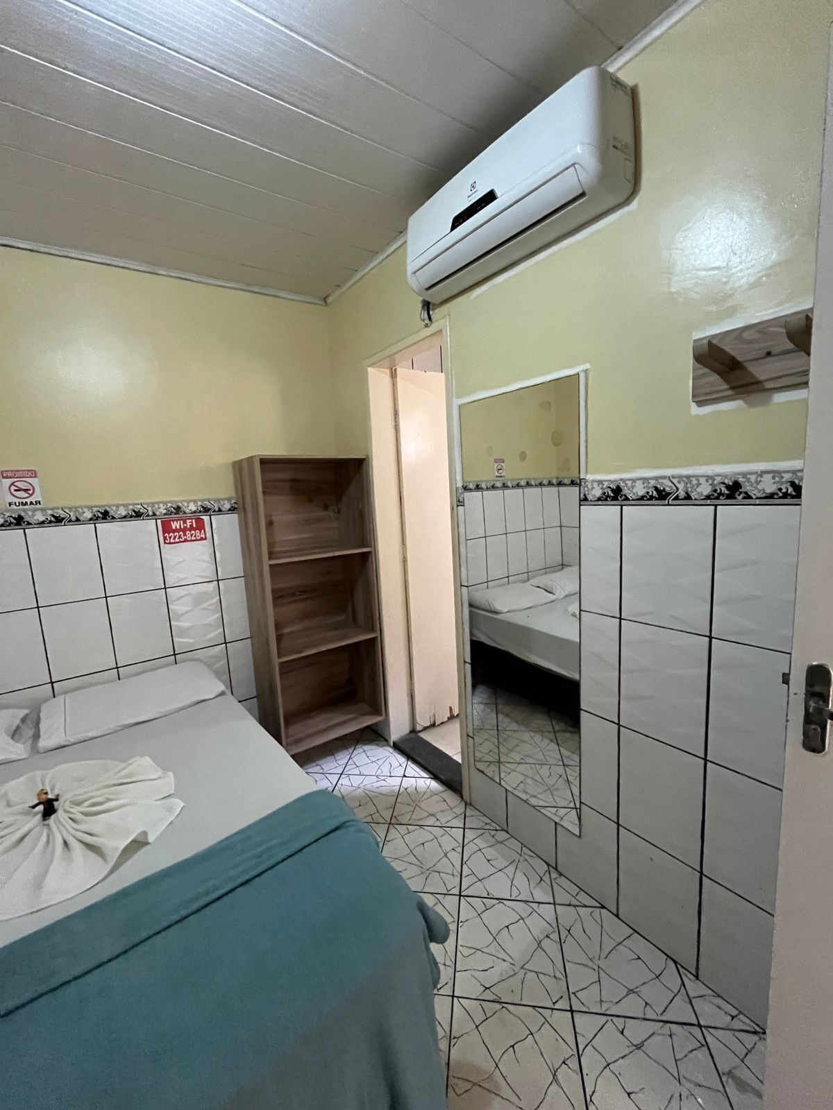 Habitación de hotel pequeña y funcional con una cama individual, un armario de madera, baño con espejo y aire acondicionado.