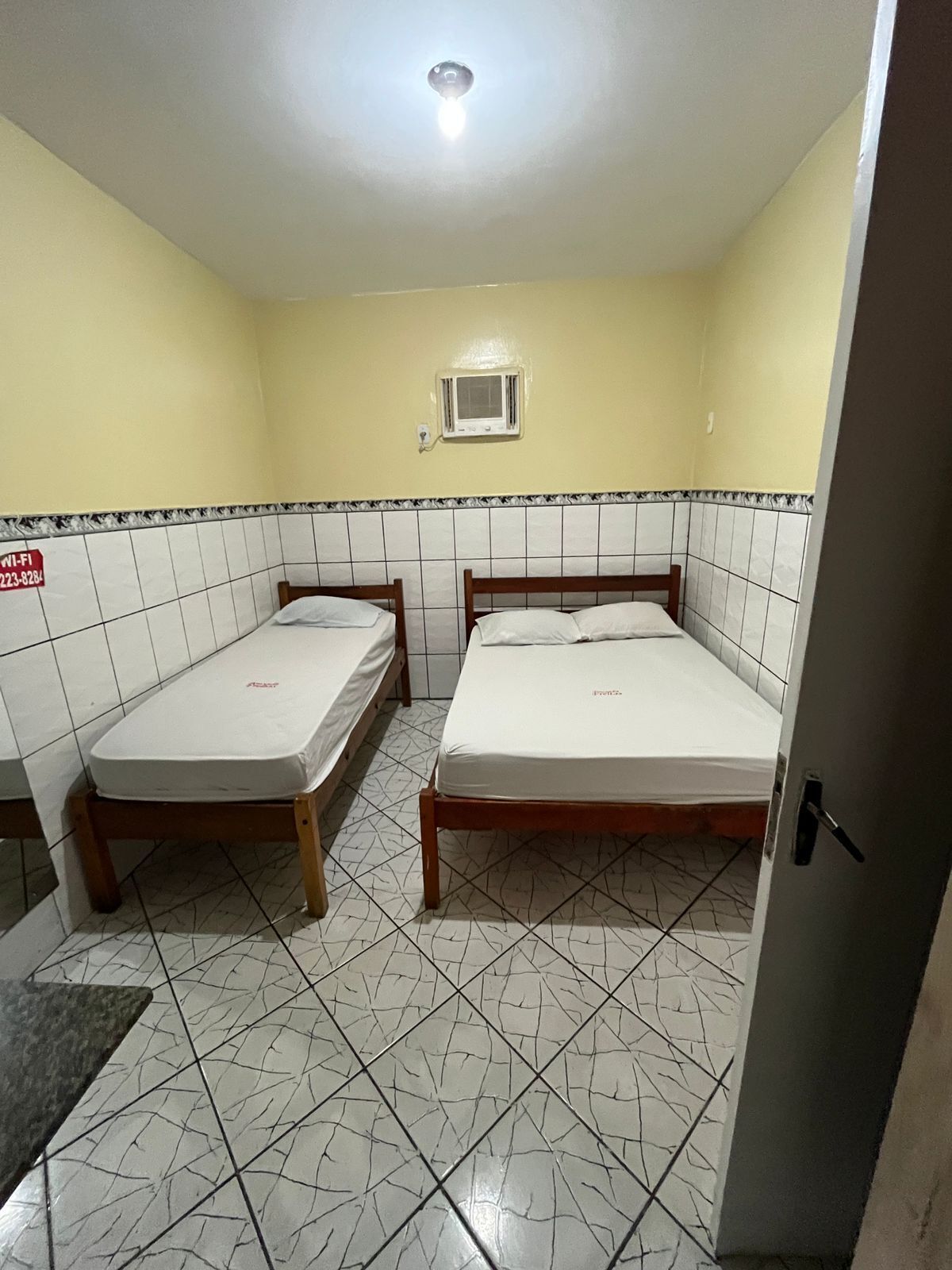 Ein kleines, sauberes Zimmer mit zwei Einzelbetten, gefliestem Boden, einer Deckenleuchte und einem kleinen Fenster mit Klimaanlage.