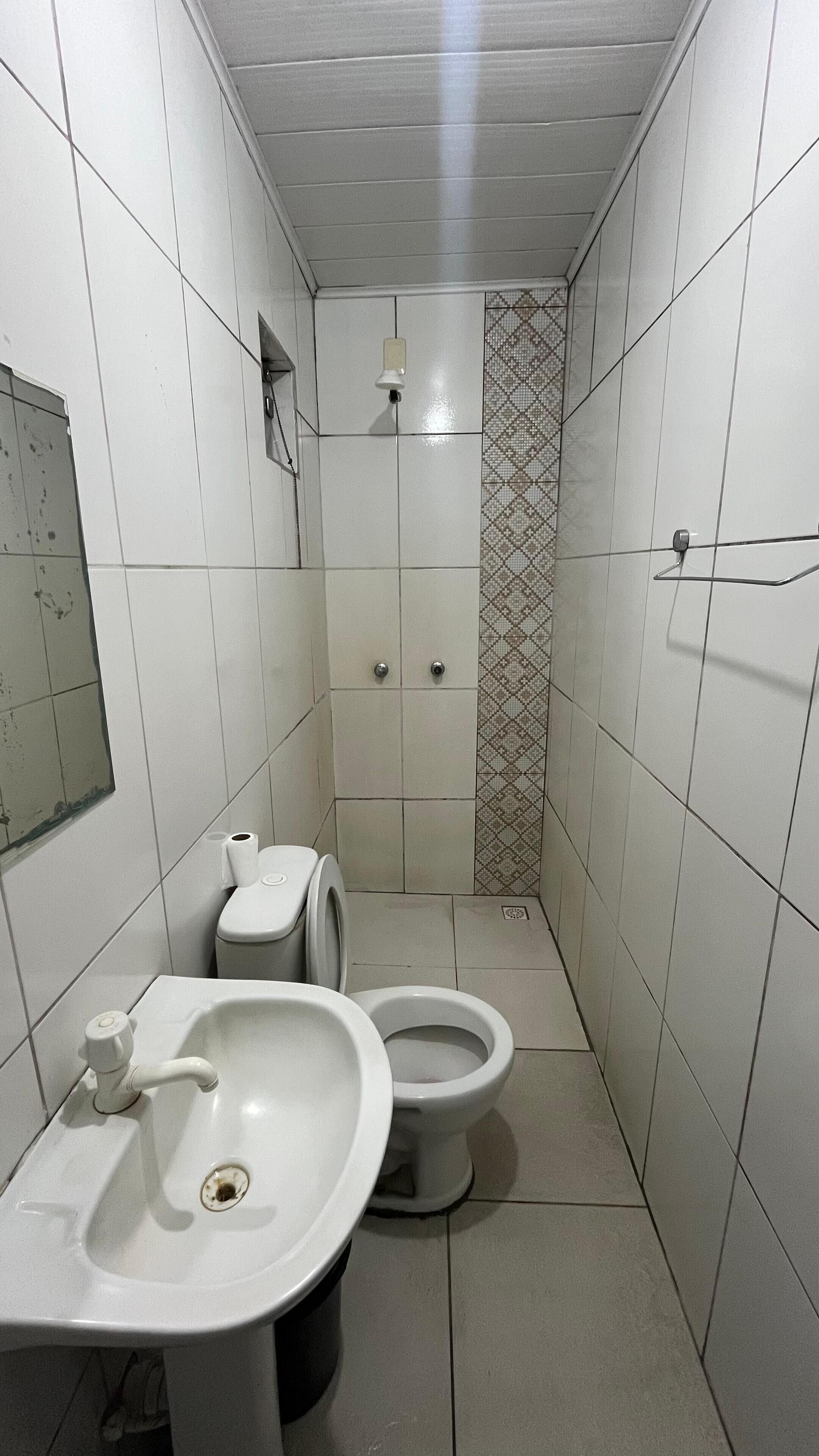 Une petite salle de bain étroite, carrelée de carreaux blancs, avec un lavabo, deux toilettes et un espace douche.