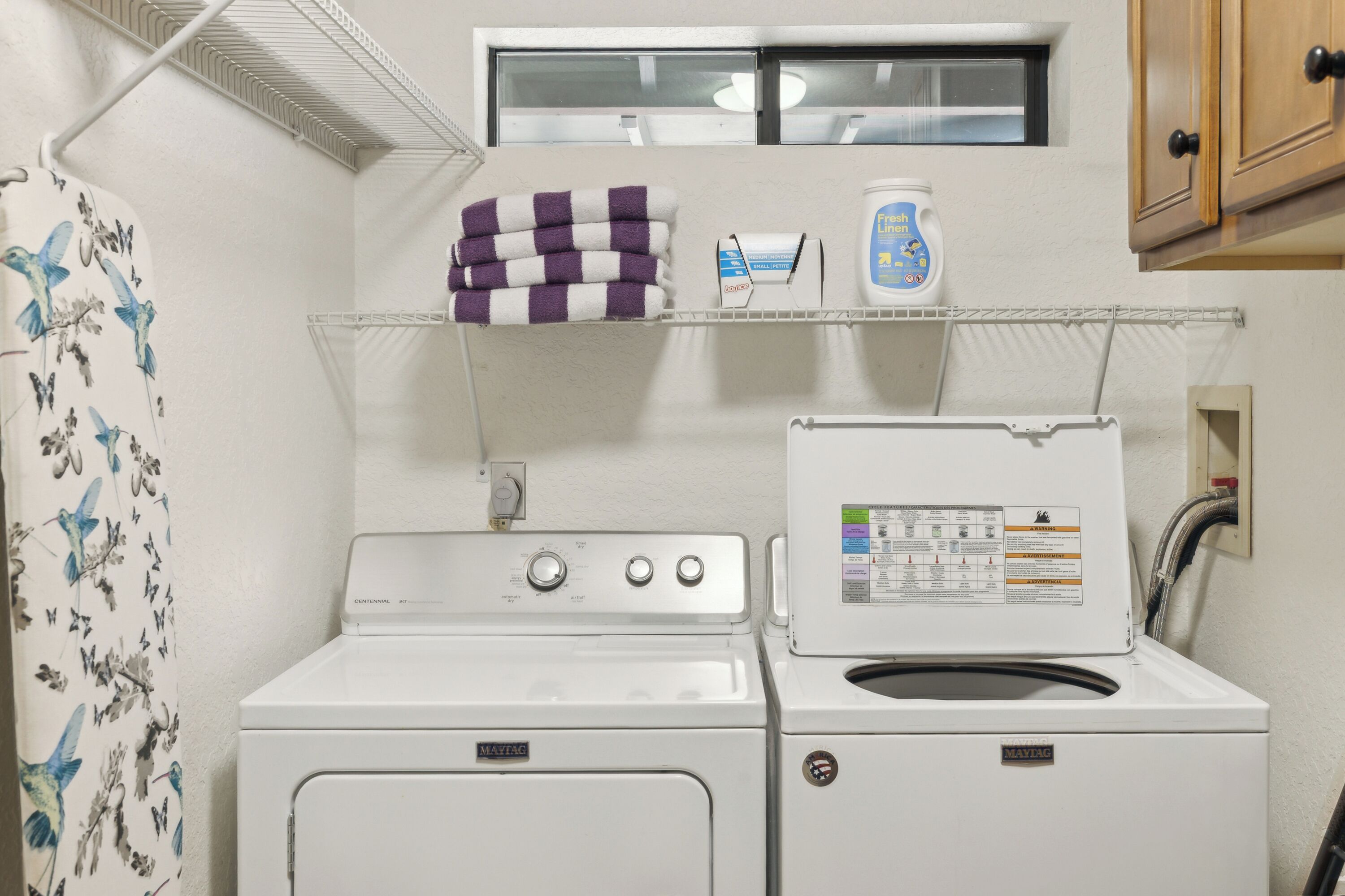 Un espace buanderie comprenant un lave-linge et un sèche-linge, une étagère avec de la lessive et une fenêtre située au-dessus du sèche-linge.