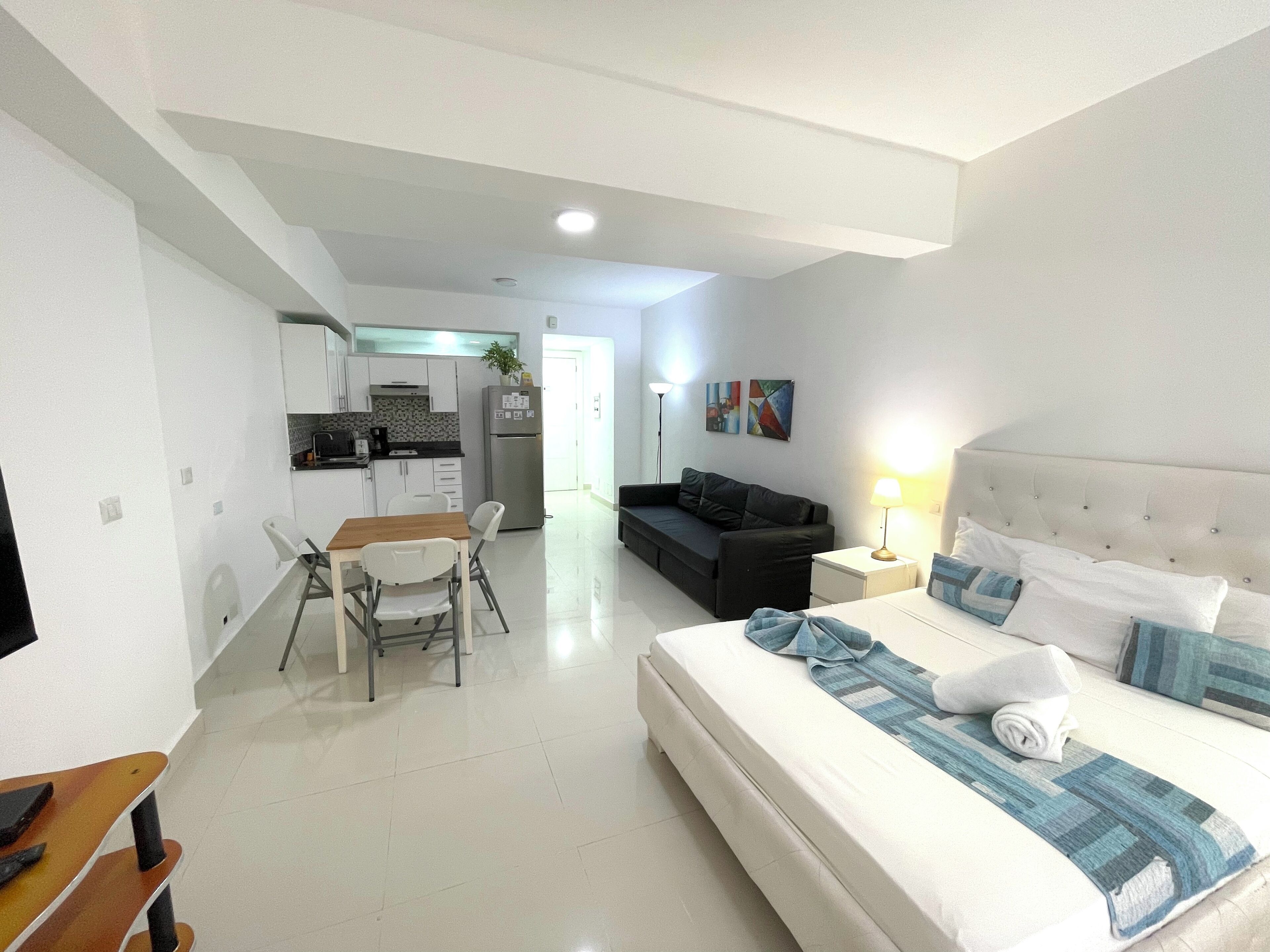 A modern hotel room with a bed, a sofa, a dining table, and a kitchen area.