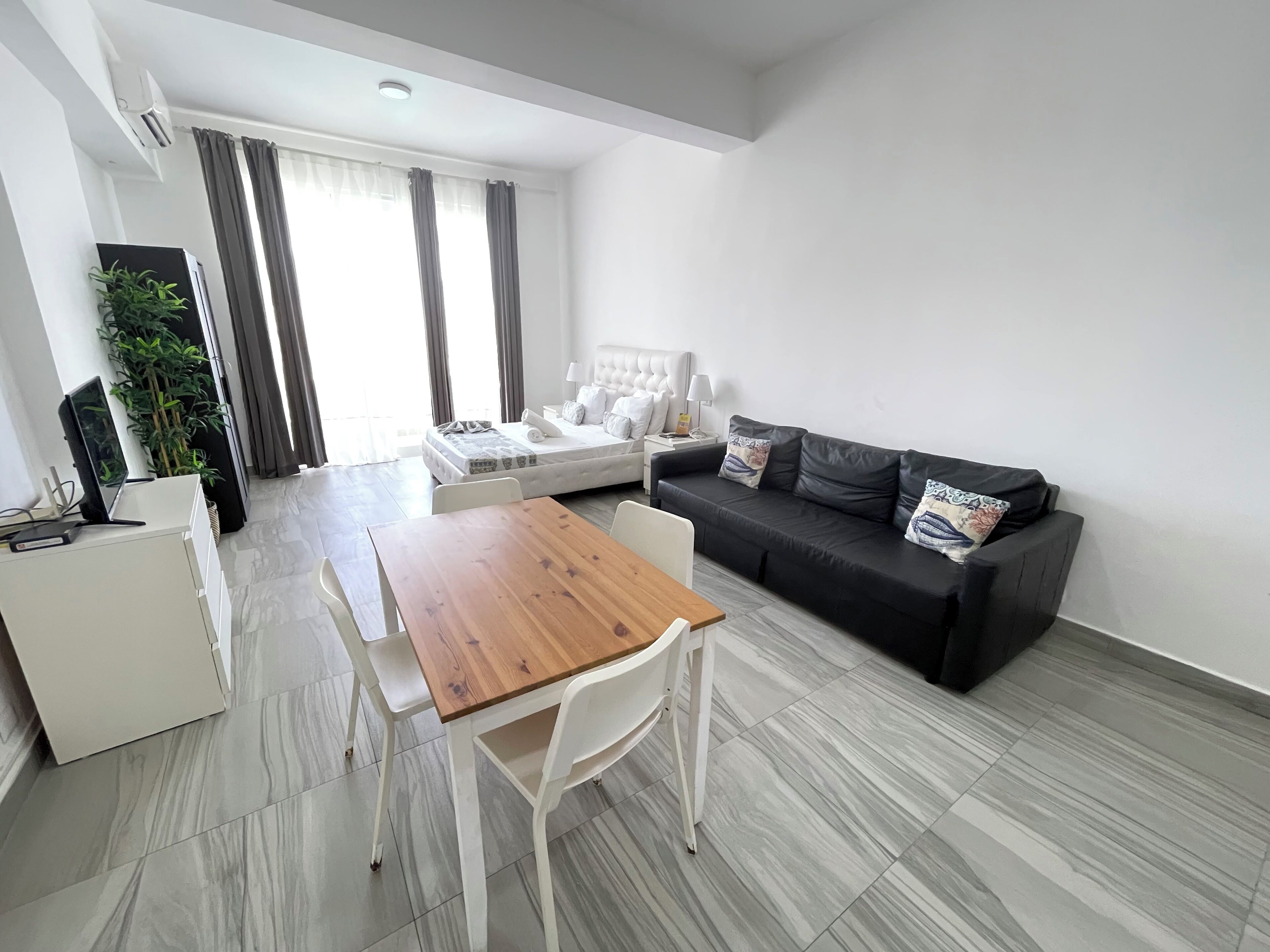 A modern living room with a wooden dining table, a black sofa, and a white bed.
