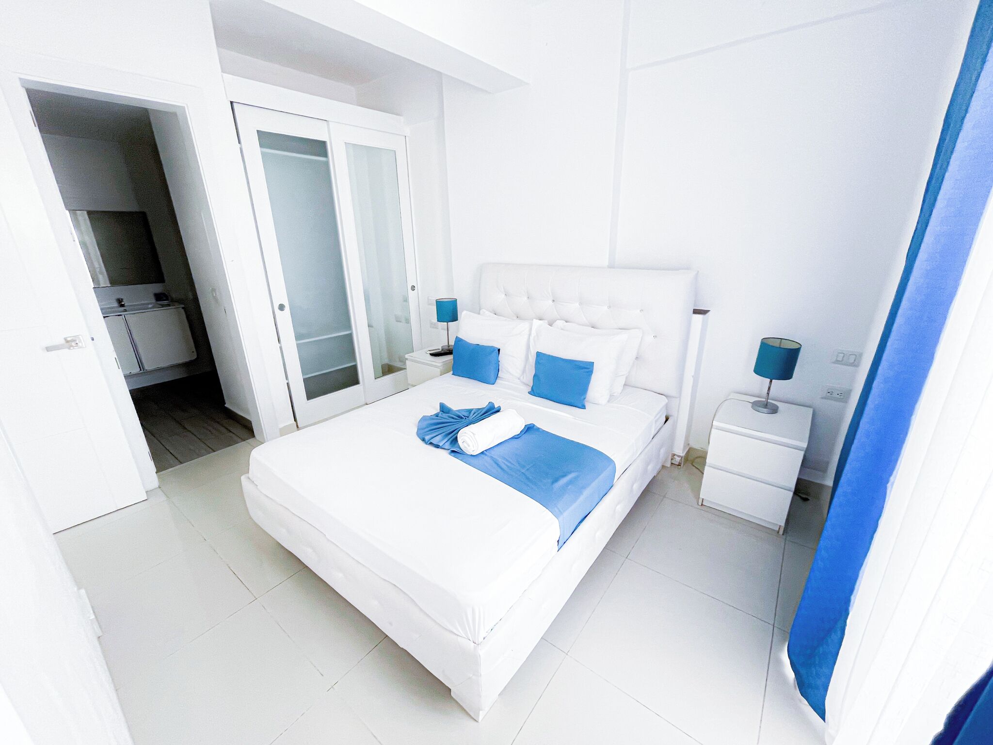 A hotel room with a large bed, a nightstand, a lamp, and a bathroom visible through an open door.