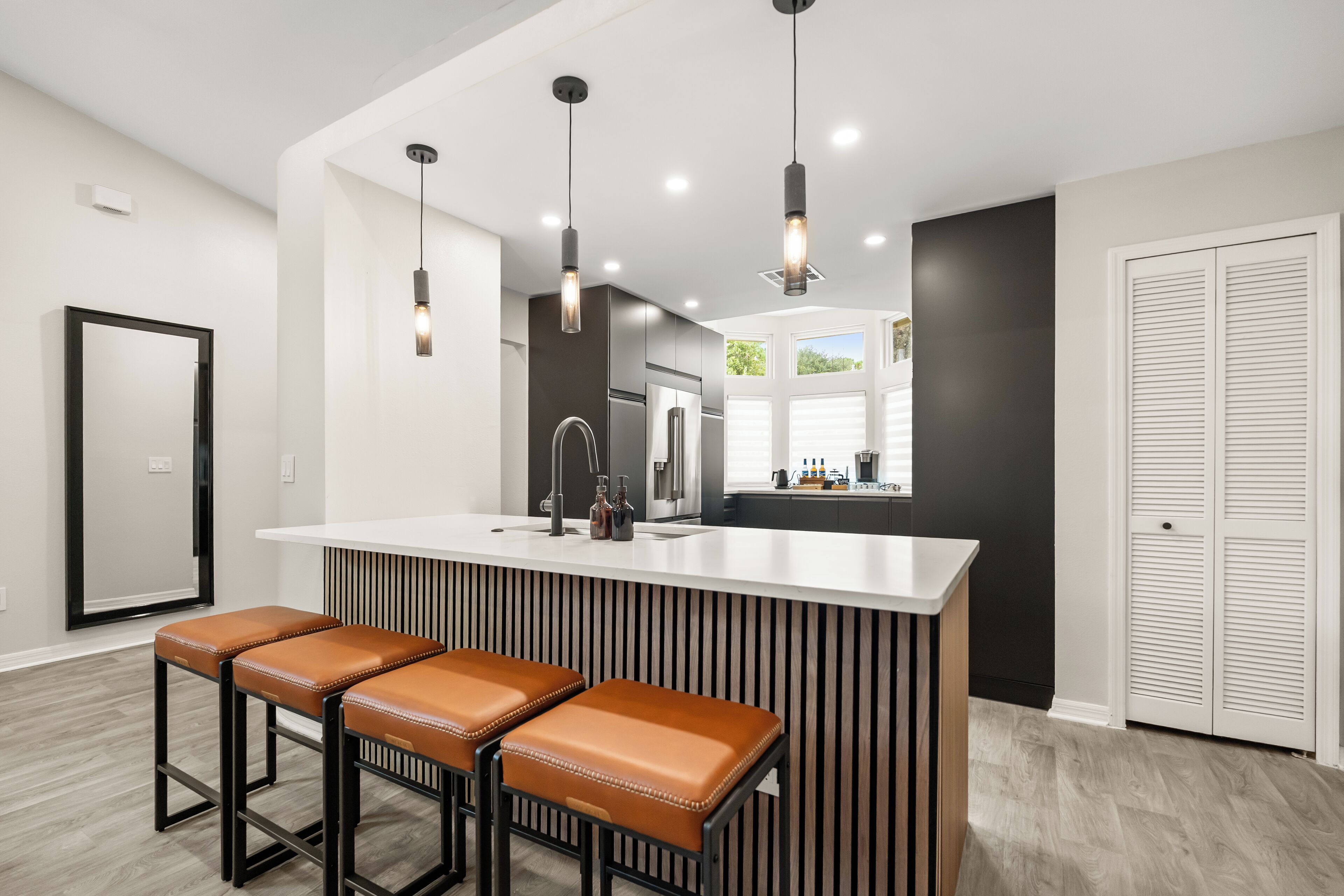 Una cuina moderna amb una illa central, tamborets de bar, il·luminació penjant i vista de la zona de dinar adjacent.