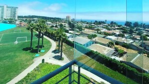 A high-rise hotel room with a balcony overlooking a residential area, a sports field, and a distant view of the ocean. - Spacious Apartment in Algarrobo in Laguna Vista with Ocean / Sea view (Algarrobo)