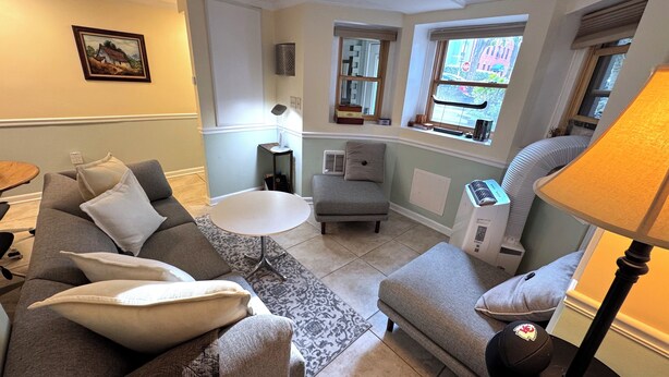 A cozy living room with a grey sofa, a small round table, a grey armchair, and a window with a view of the street.