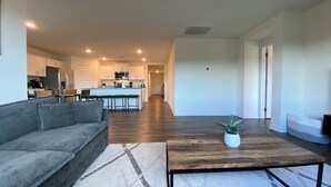 Amplia sala de estar con un sofá gris, una mesa de centro de madera y una cocina al fondo.