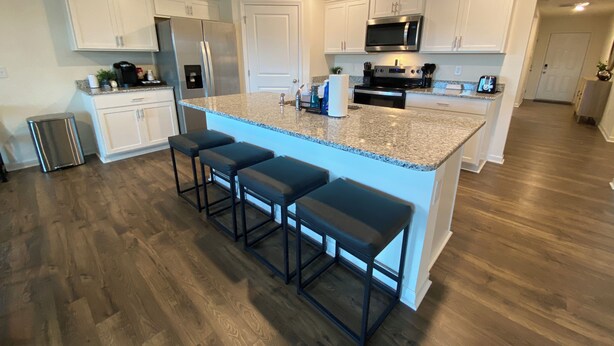 Una cocina moderna con armarios blancos, encimera de granito, electrodomésticos de acero inoxidable y una isla central con taburetes de bar.