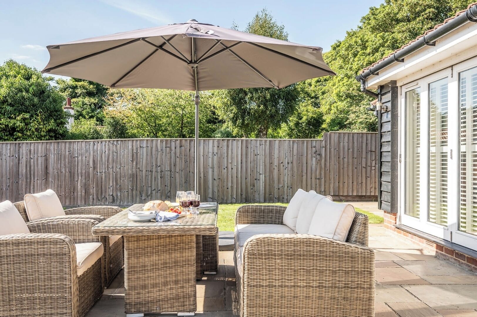 Une terrasse extérieure aménagée avec des meubles en osier, une table dressée avec des assiettes et des verres, et un grand parasol.