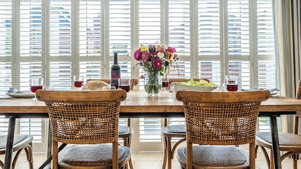 Un coin repas avec une table en bois, des chaises en osier, un vase de fleurs et une bouteille de vin.