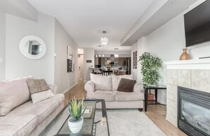 A modern living room with a sofa, coffee table, and fireplace. There is a dining area in the background with a chandelier.