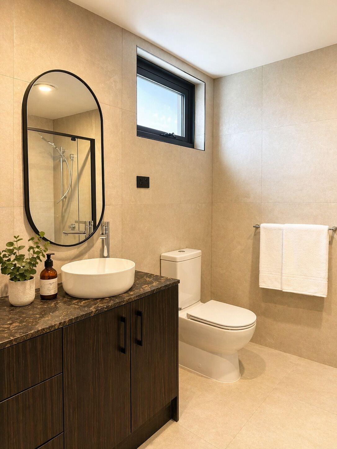 A modern bathroom with a large oval mirror, a vessel sink, a toilet, and a window above the sink.