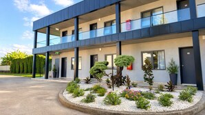 Een modern meerlaags gebouw met een plat dak, grote ramen en een aangelegde tuin met diverse planten en een grindpad.