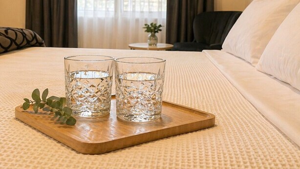 Hotelkamer met een bed, twee glazen op een dienblad, een kleine tafel met een vaas en uitzicht door een raam met gordijnen.