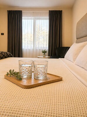 A hotel room with a bed, two glasses on a tray, a small table with a vase, and a view through a window with curtains. - Jolly Knight Motel (Casula)