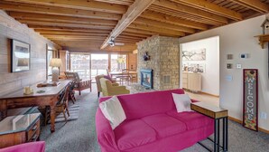 Un intérieur de chalet chaleureux avec une cheminée en pierre, des poutres en bois, un canapé rose et un coin repas offrant une vue sur l’extérieur.