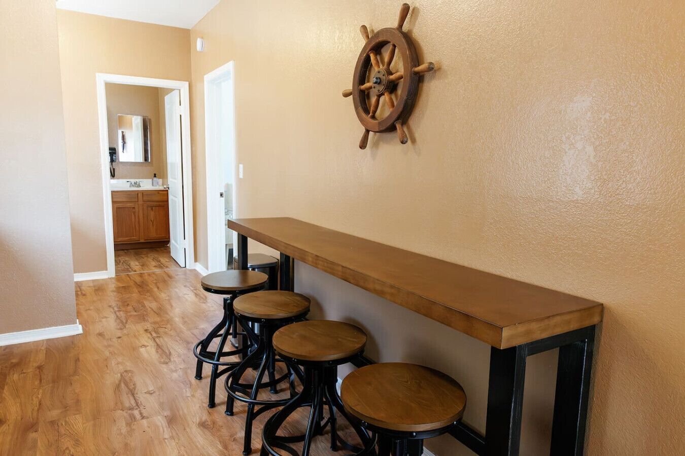 Un couloir avec un banc et des tabourets en bois, une décoration représentant un gouvernail de navire au mur, et un coin cuisine à l’arrière-plan.