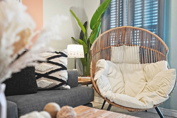 A wicker chair with cushions, a grey sofa, a lamp, and a large plant in a living room.