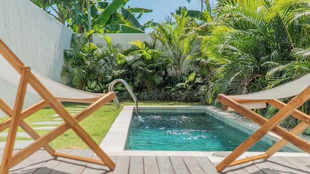 A pool area with two wooden lounge chairs, surrounded by lush tropical plants and a clear blue sky. - Sunny Villa Tumbak Bayuh (Canggu)