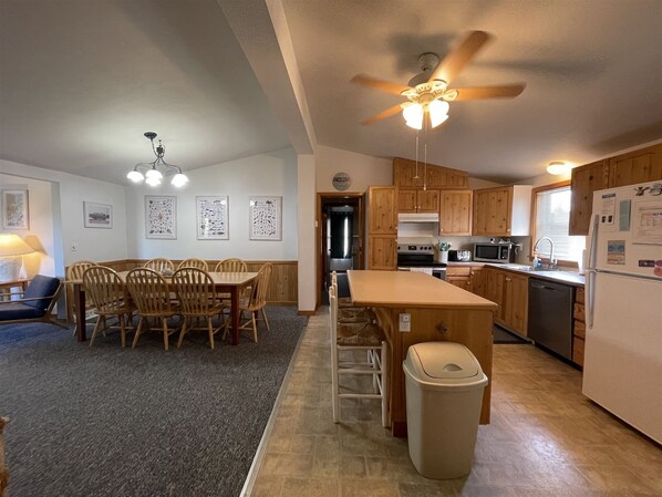 A compact kitchen with wooden cabinets, a central island, a ceiling fan, and a dining area with chairs and a table. - Chasing Sunsets Dog-friendly home w/deck and ocean view (HATTERAS)