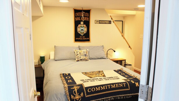 Una cama bien hecha con una manta de color azul marino y estampado dorado, un cartel en la pared que dice "United States Naval Academy" y una mesita de noche con una lámpara al lado de la cama.