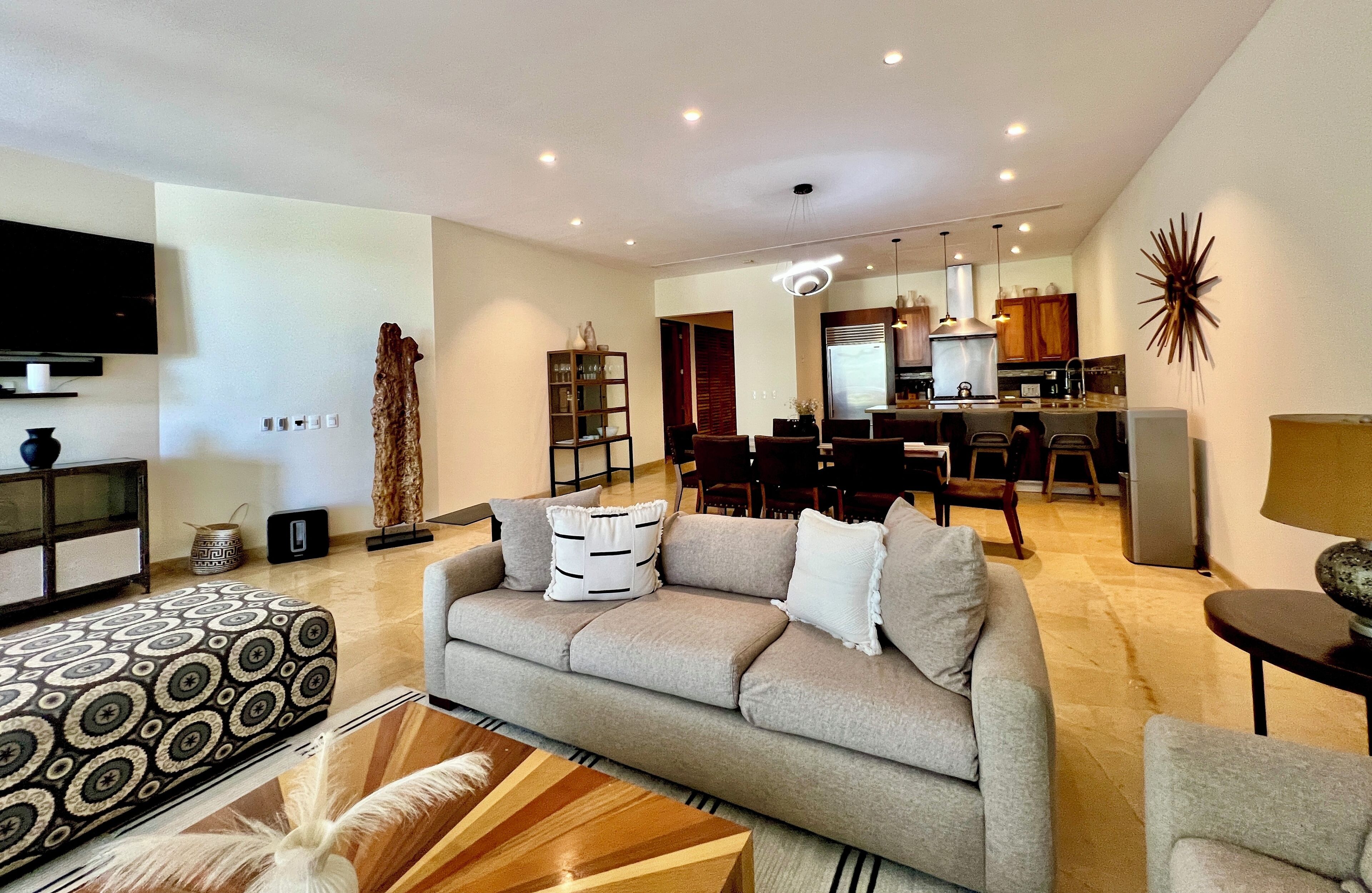 A modern living room with a sofa, coffee table, and TV. There is a dining area with chairs and a table. The kitchen is visible in the background with cabinets and appliances.