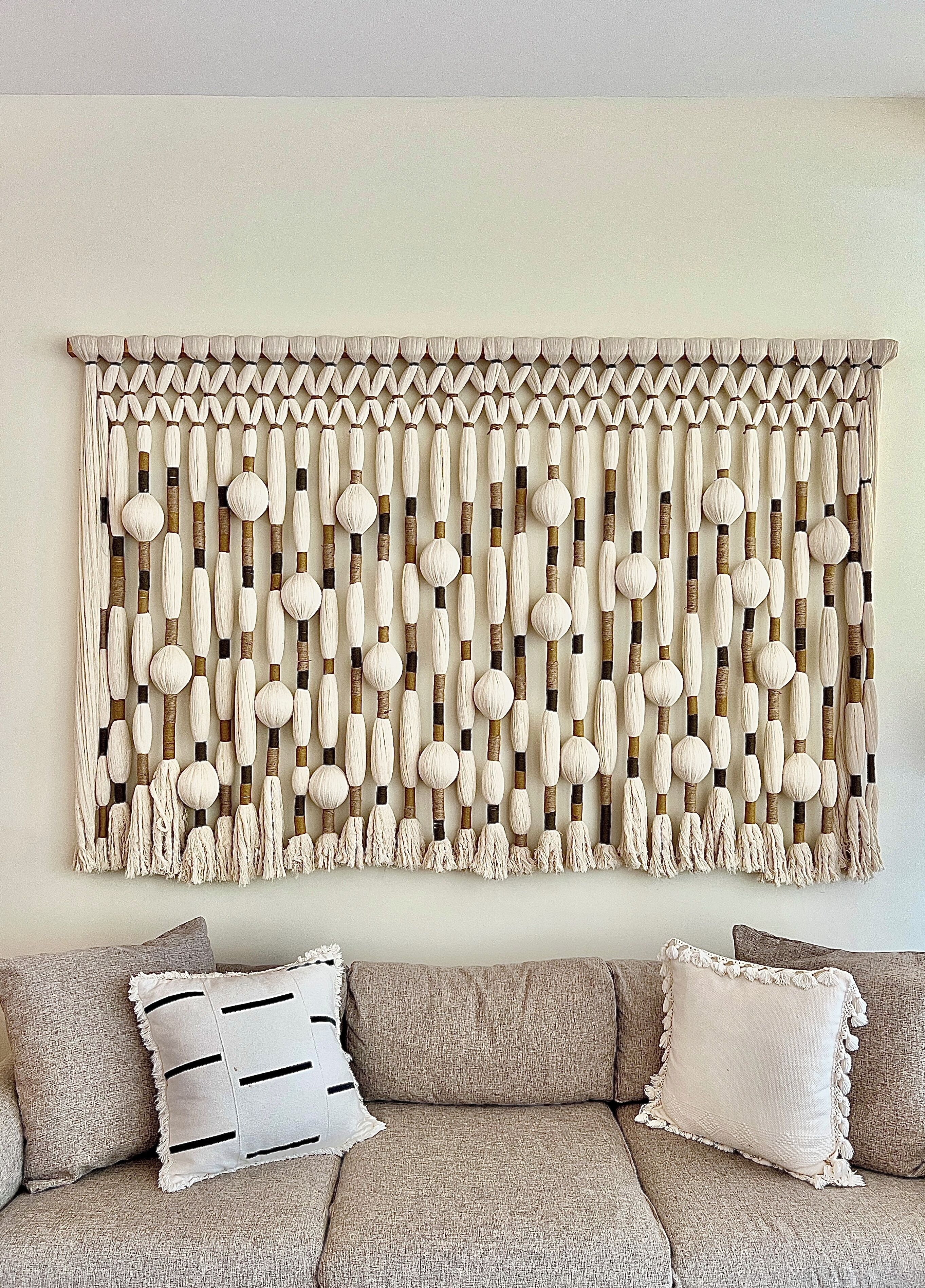 Une œuvre en macramé fixée au mur, composée d’éléments sphériques et cylindriques, un canapé à texture intéressante agrémenté de coussins à motifs, et un mur aux tons neutres.