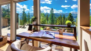 Ein Zimmer mit großen Fenstern und Blick auf die Berge, ein Holztisch mit einem Buch und einer Topfpflanze.