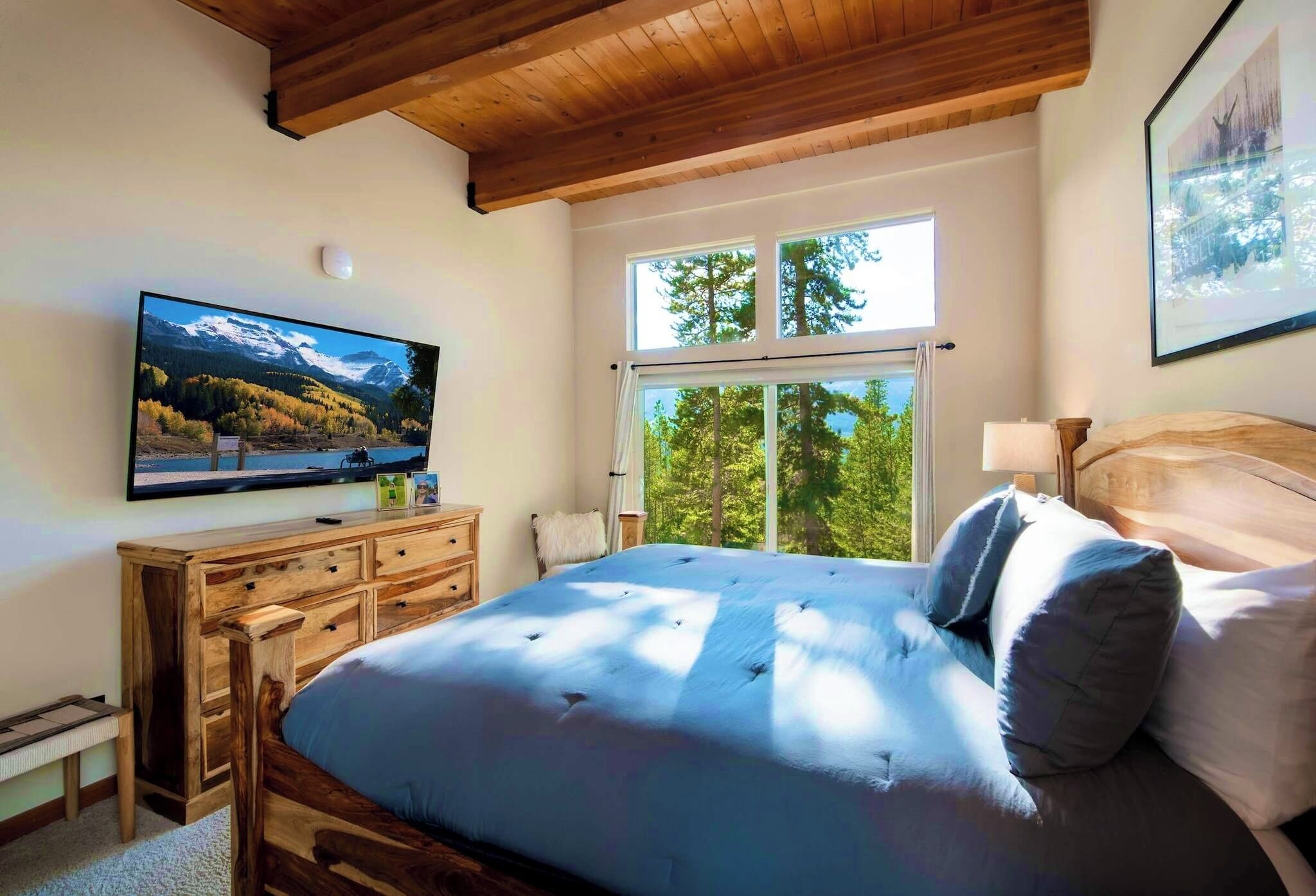 Une chambre avec un lit en bois, un téléviseur fixé au mur et une fenêtre donnant sur des arbres.