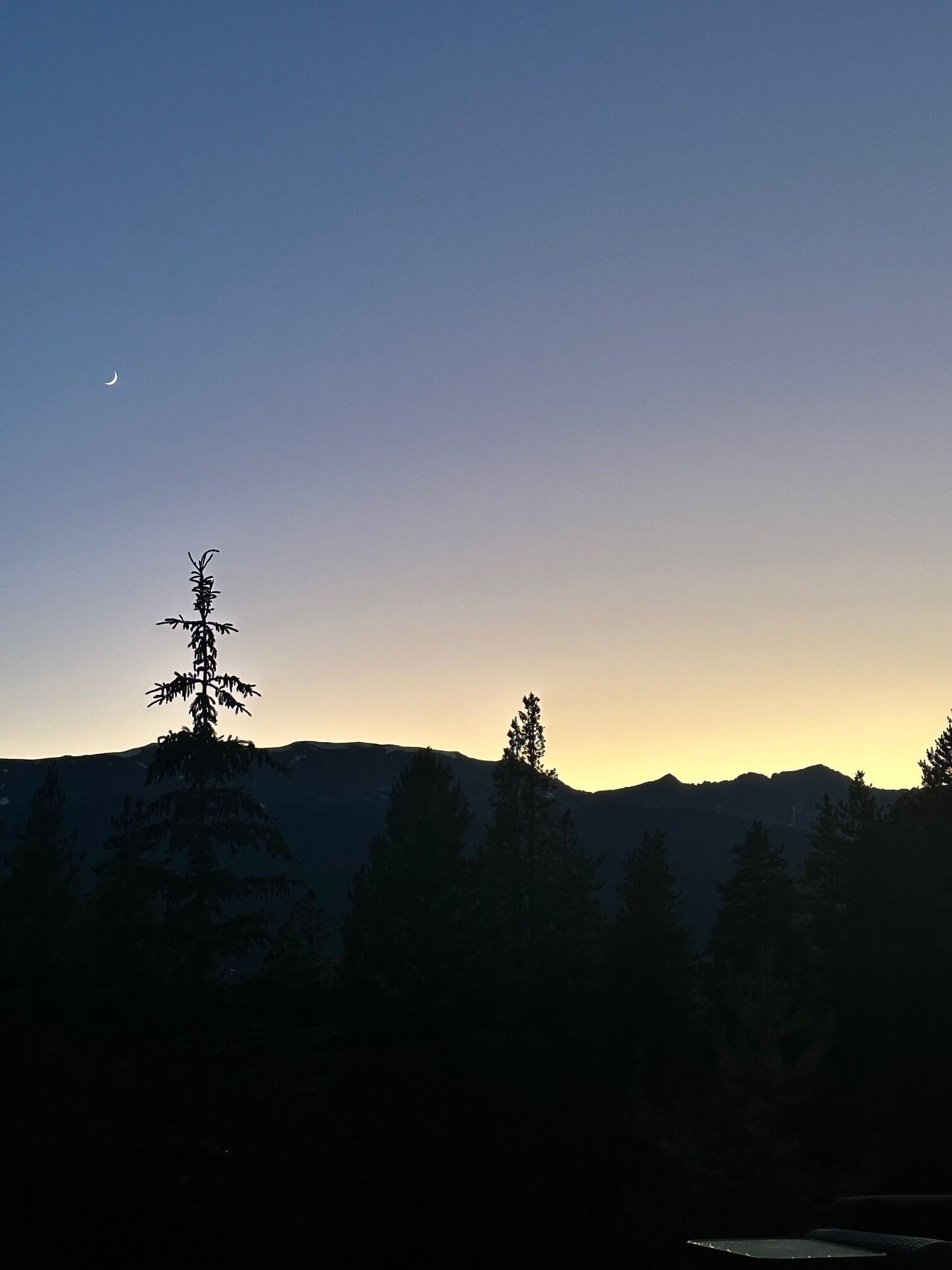 Un paysage de montagne avec un croissant de lune dans le ciel et les silhouettes d’arbres.