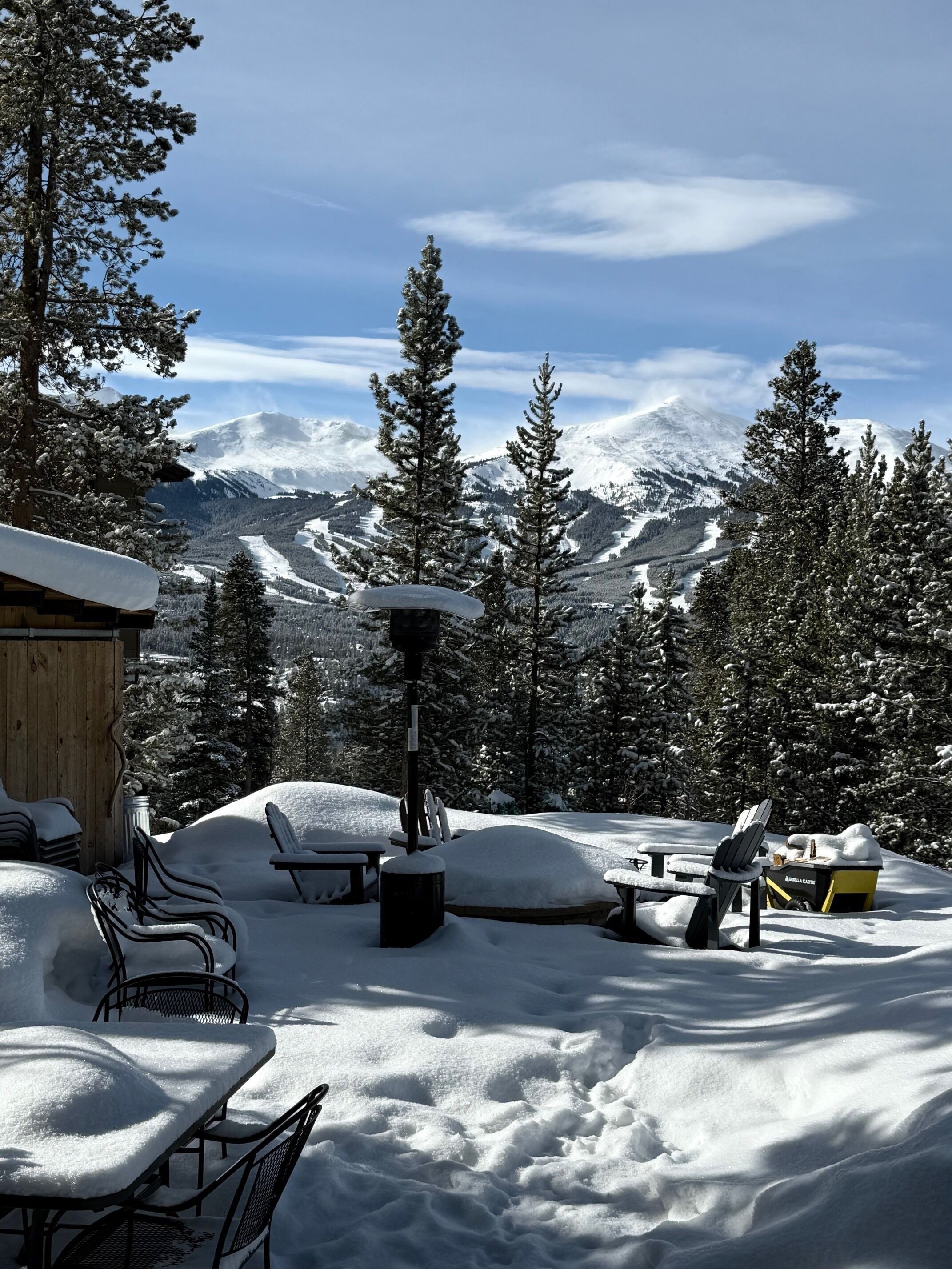 A snow-covered outdoor seating area with chairs and tables, surrounded by snow-laden trees and mountains in the background.