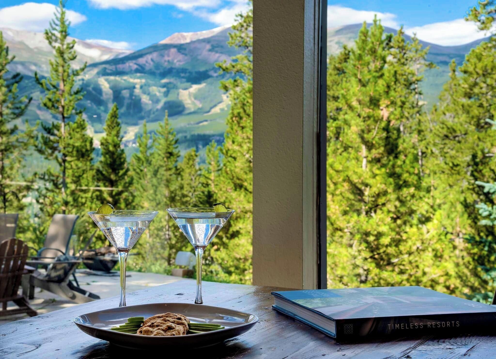 Une table avec deux verres à martini, une assiette de pains plats et un livre intitulé « Les plus beaux hôtels de tous les temps », le tout sur fond de paysage montagneux.