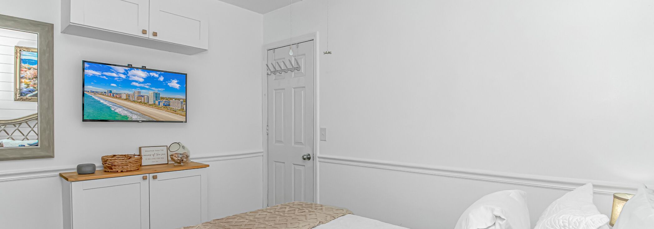 A bedroom with a bed, a TV mounted on the wall, a door, and a small shelf with items.