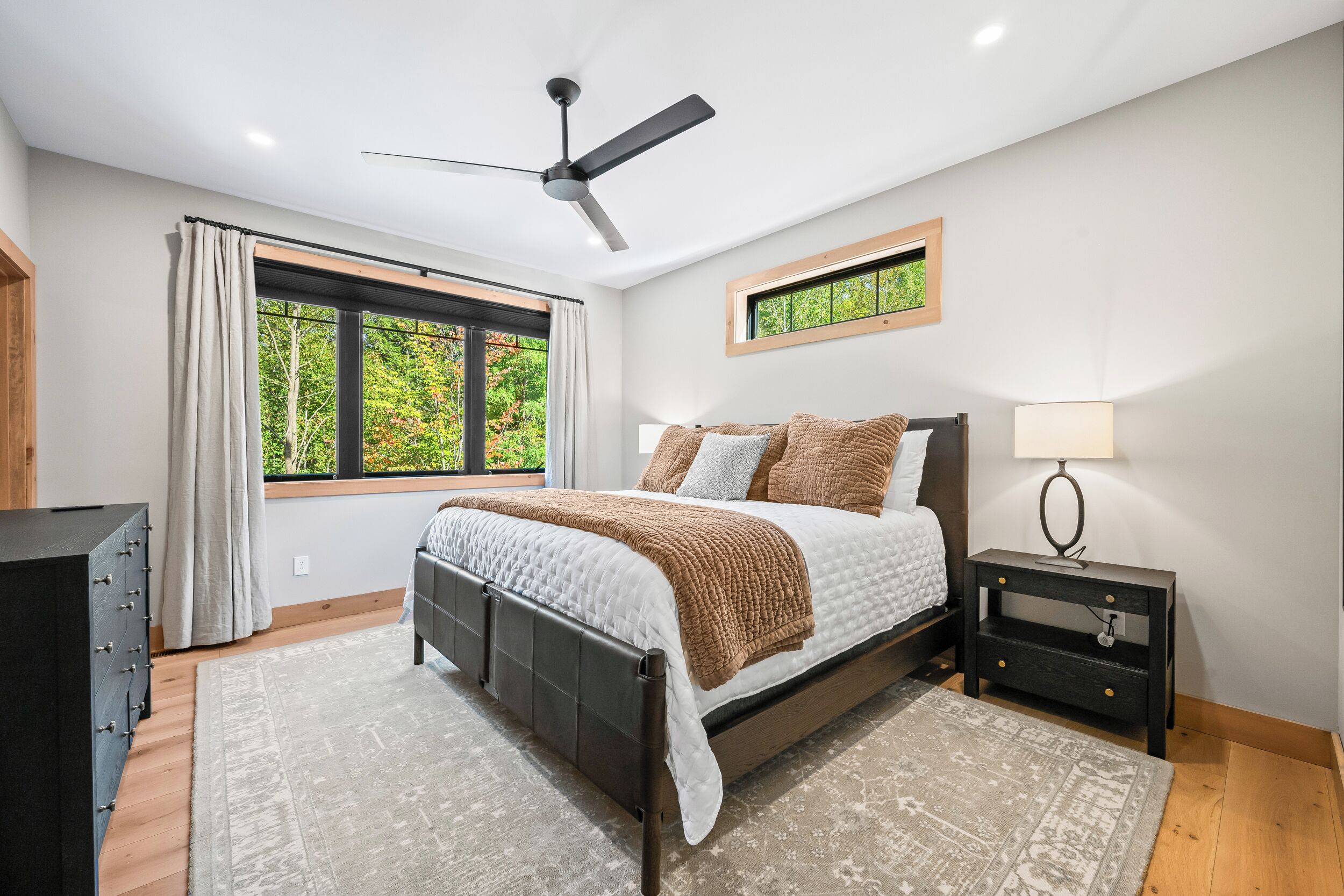 A bedroom with a bed, a ceiling fan, a nightstand, a window with a view of trees, and a wooden floor.