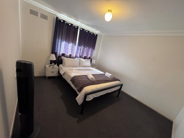 A neatly made bed with white linens, a dark brown throw blanket, and a rolled towel on the footboard. There is a white bedside table with a lamp and a small decorative item. The room has a dark carpet, white walls, and a window with dark curtains.