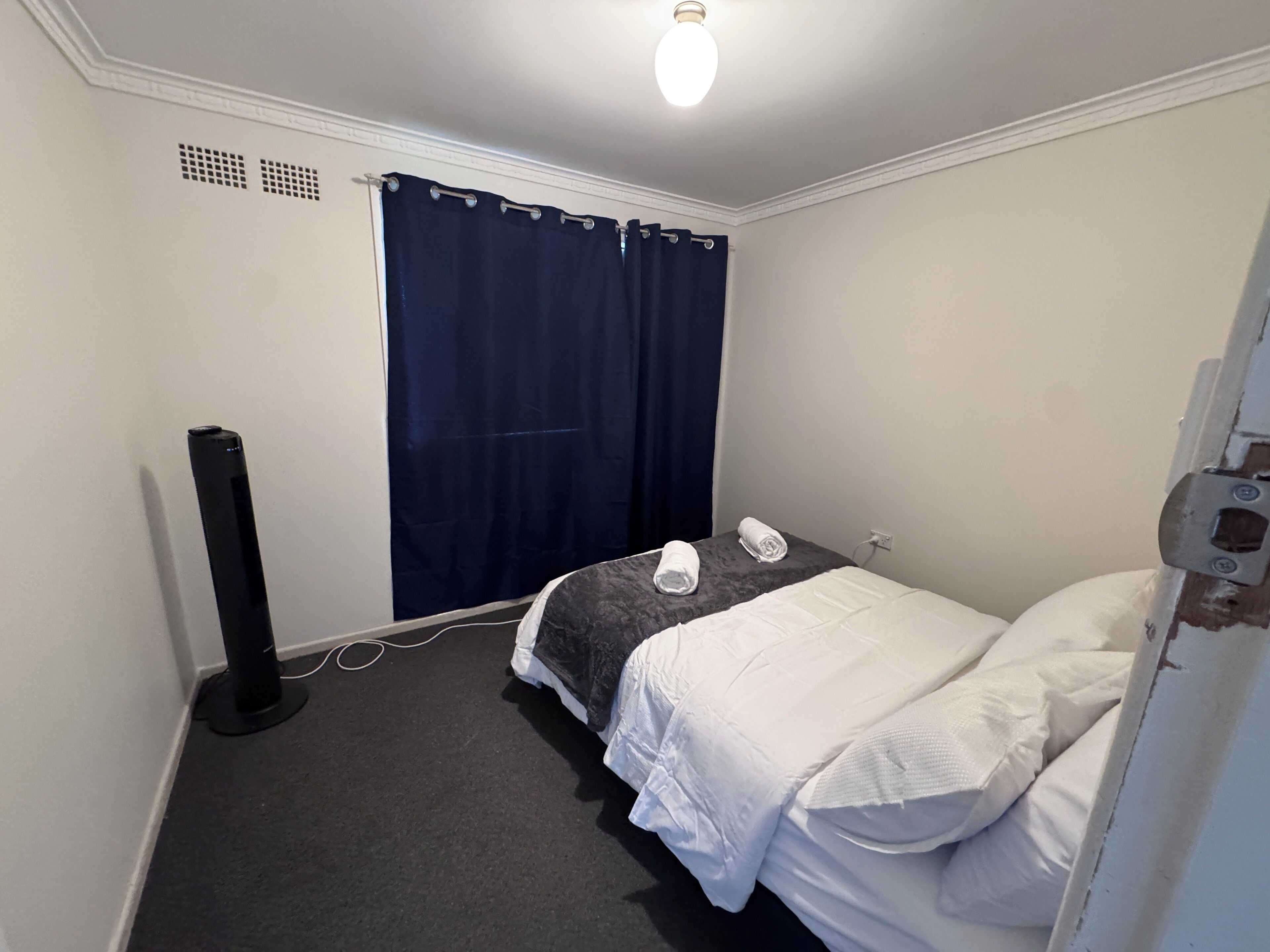 Una habitación pequeña y limpia con cama, escritorio y silla. Cuenta con una ventana con cortinas, un calentador instalado en la pared y una puerta con manija.
