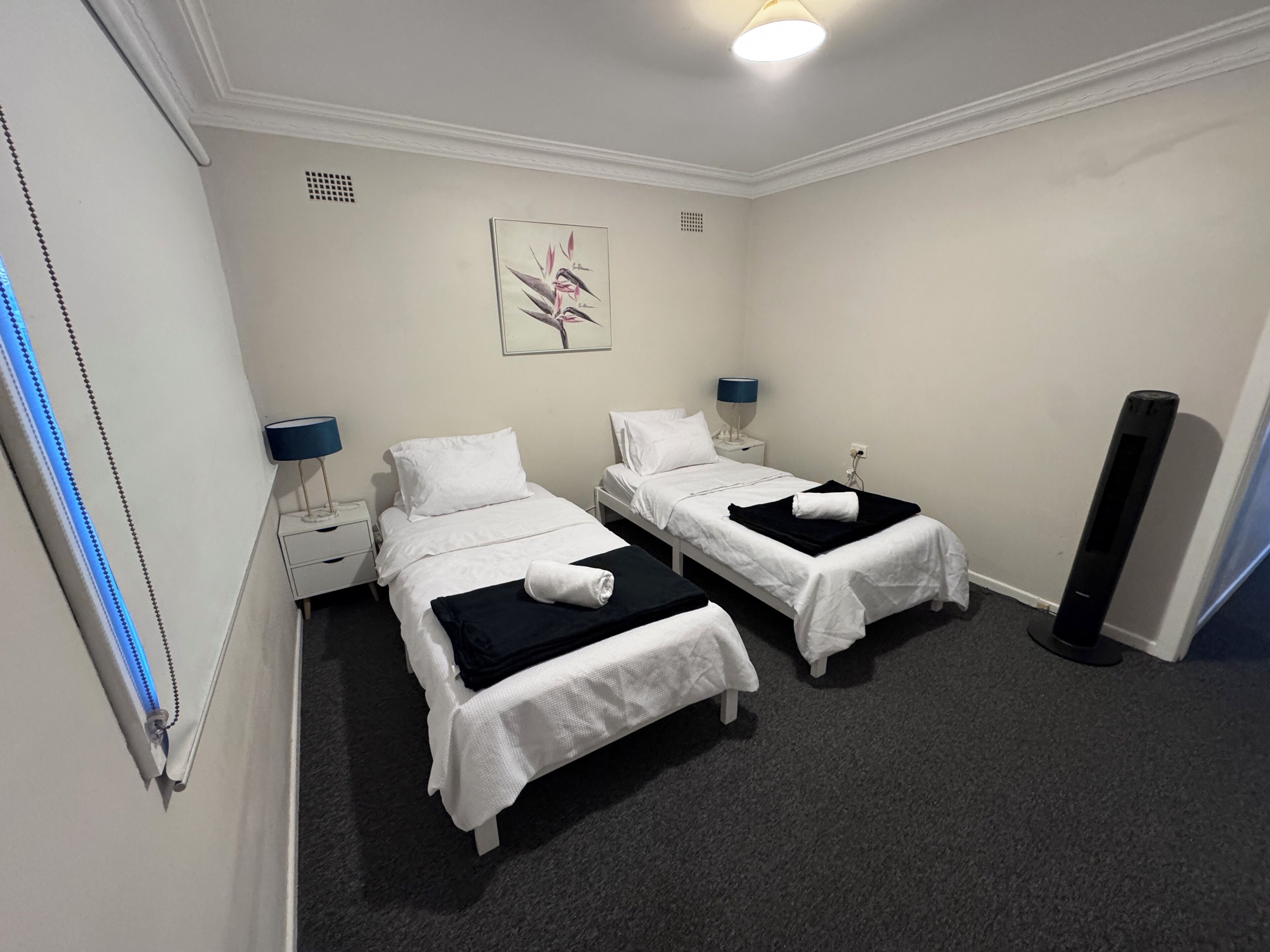 A hotel room with two single beds, a nightstand with a lamp, a wall-mounted artwork, and a standing heater.