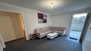 Un salon avec un canapé d’angle beige, une table basse blanche et une petite table d’appoint. Une porte donne accès à un balcon avec vue sur l’extérieur.