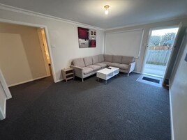 Una sala de estar con un sofá seccional color beige, una mesa de centro blanca y una mesita auxiliar. Hay una puerta que da a un balcón con vistas al exterior.