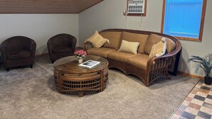 Un salon chaleureux avec un canapé en osier marron, deux fauteuils, une table basse ronde en osier agrémentée d’un livre et de fleurs, un ventilateur de plafond, une fenêtre avec des stores et un climatiseur fixé au mur.
