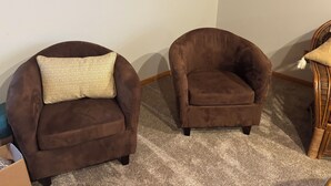 Deux fauteuils marron, dotés de coussins à motifs, se trouvent dans une pièce dont le plafond est en bois et le sol recouvert d’une moquette.