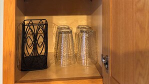 Un meuble en bois avec trois étagères ; l’étagère du haut contient un sac en plastique, celle du milieu est remplie d’assiettes empilées, et l’étagère du bas abrite un panier en fil métallique noir et trois verres transparents.