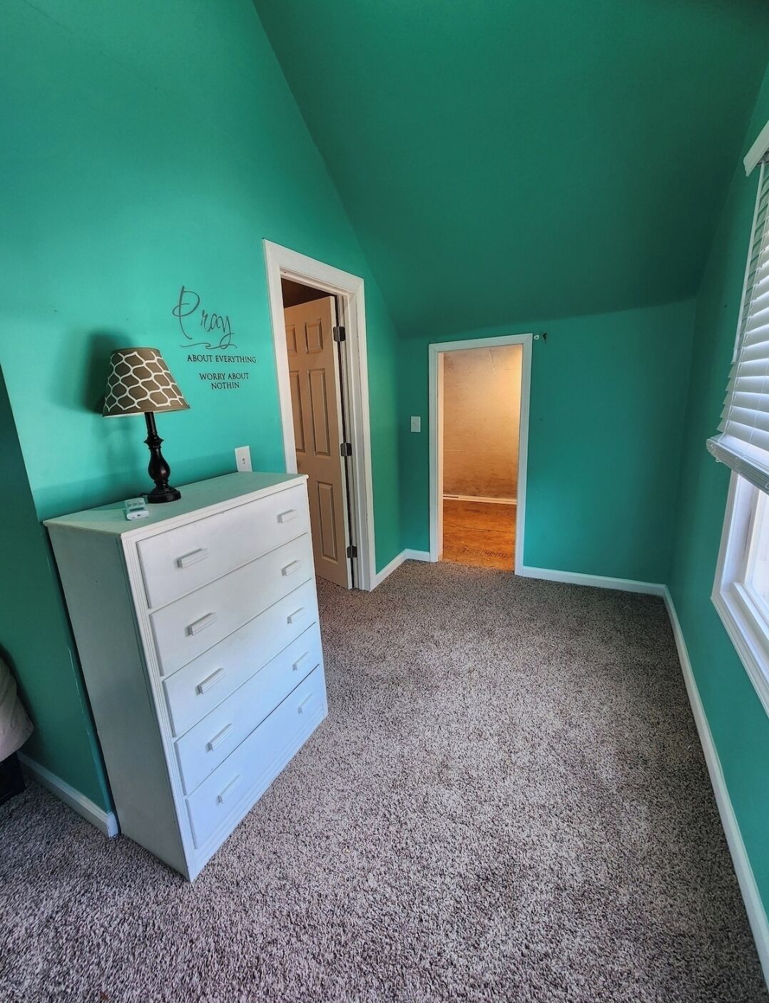 Una habitación con paredes verde azulado, un tocador blanco, una lámpara y una puerta que da a otra habitación.