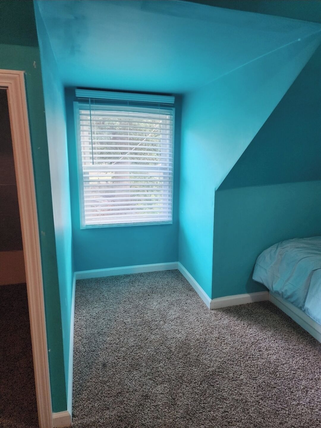 Een kleine, kamer-en-zolder slaapkamer met blauwgroene wanden, een raam met zonwering en een vloerbedekking.